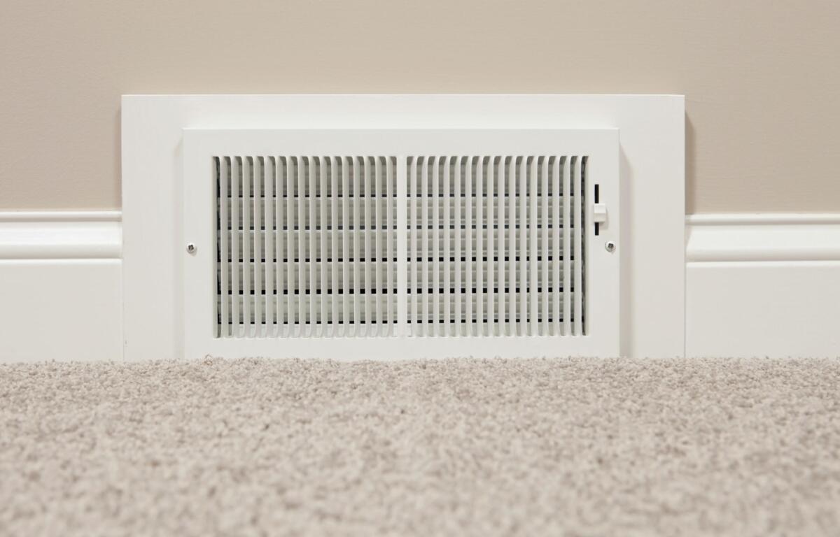 White rectangular wall vent with vertical slats set into a beige wall above light carpet, part of an HVAC system.