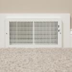 White rectangular wall vent with vertical slats set into a beige wall above light carpet, part of an HVAC system.