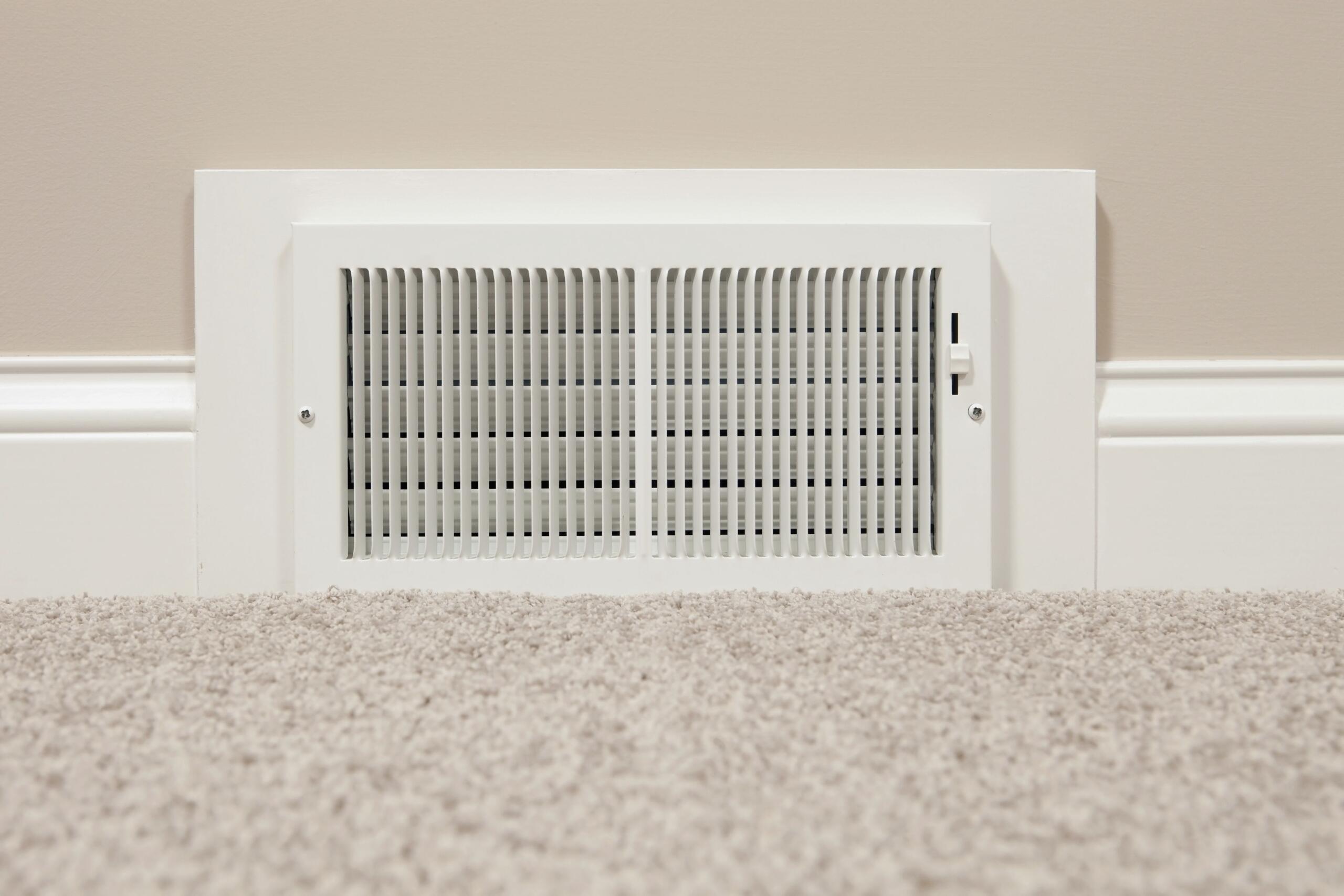 White rectangular wall vent with vertical slats set into a beige wall above light carpet, part of an HVAC system.