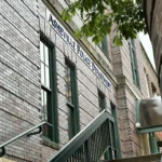 Brick building exterior with green-framed windows and an exterior metal stair railing along a narrow alley. (Informative)