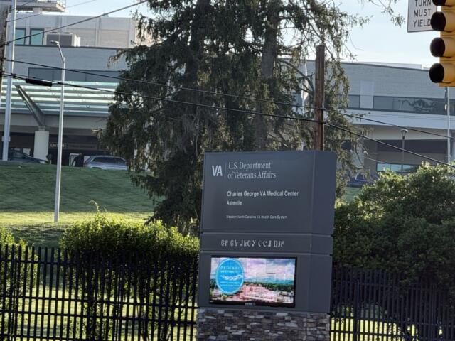 VA sign for the U.S. Department of Veterans Affairs with Charles George VA Medical Center, Asheville, visible beside a fence and greenery.