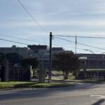 Urban street scene with power lines and a tall utility pole, a modern gray building in the background, and a fenced grassy area in the foreground.