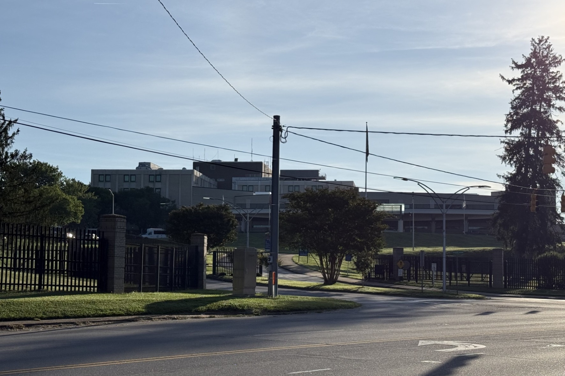 Urban street scene with power lines and a tall utility pole, a modern gray building in the background, and a fenced grassy area in the foreground.