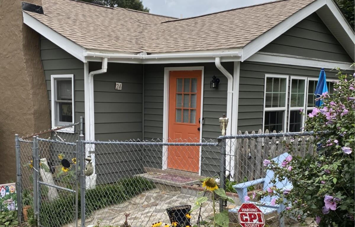 Gray siding house with an orange front door, white trim, and a fenced front yard with sunflowers and blooming pink flowers on the right term garden.
