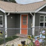 Gray siding house with an orange front door, white trim, and a fenced front yard with sunflowers and blooming pink flowers on the right term garden.