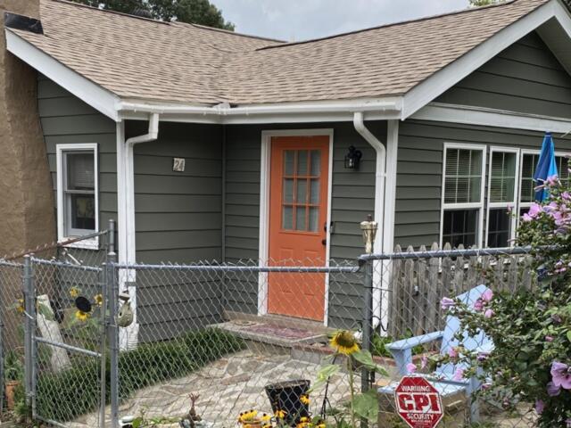 Gray siding house with an orange front door, white trim, and a fenced front yard with sunflowers and blooming pink flowers on the right term garden.