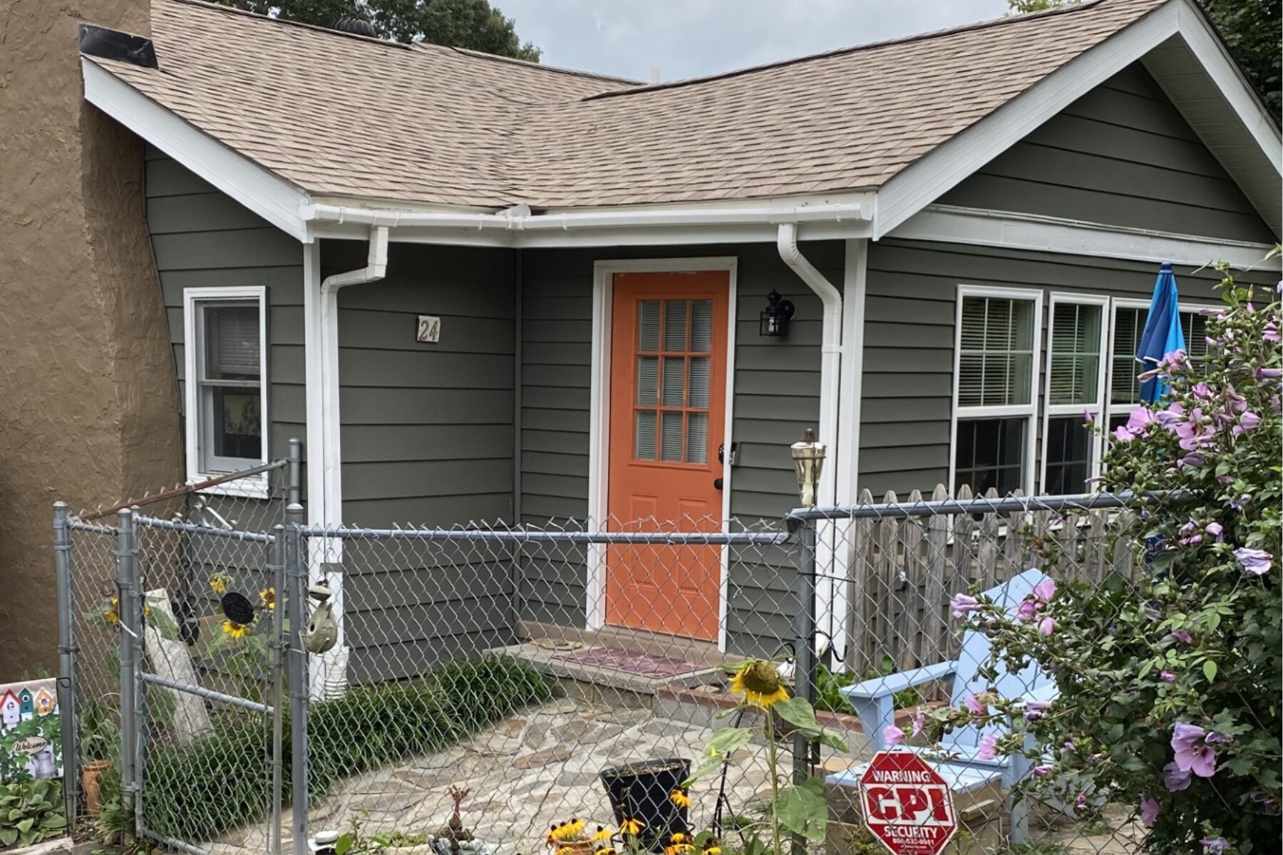 Gray siding house with an orange front door, white trim, and a fenced front yard with sunflowers and blooming pink flowers on the right term garden.