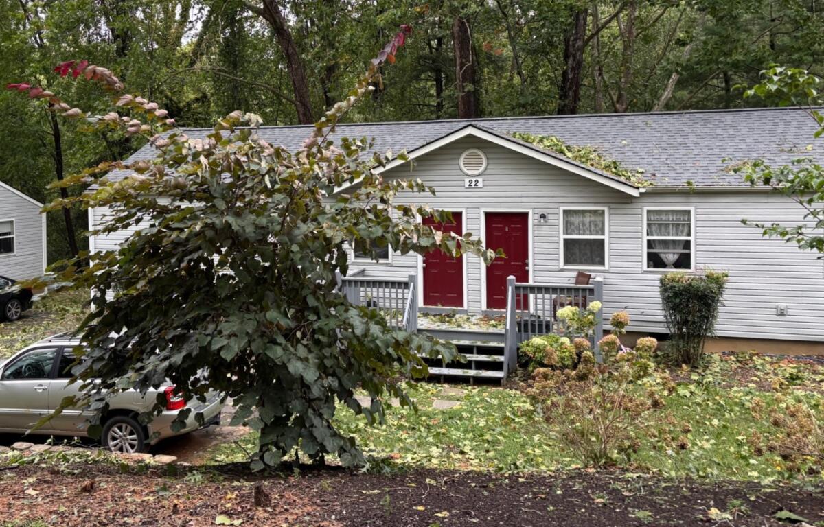 Two red doors on a gray siding duplex with a small front porch, surrounded by trees and parked cars nearby.