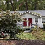 Two red doors on a gray siding duplex with a small front porch, surrounded by trees and parked cars nearby.