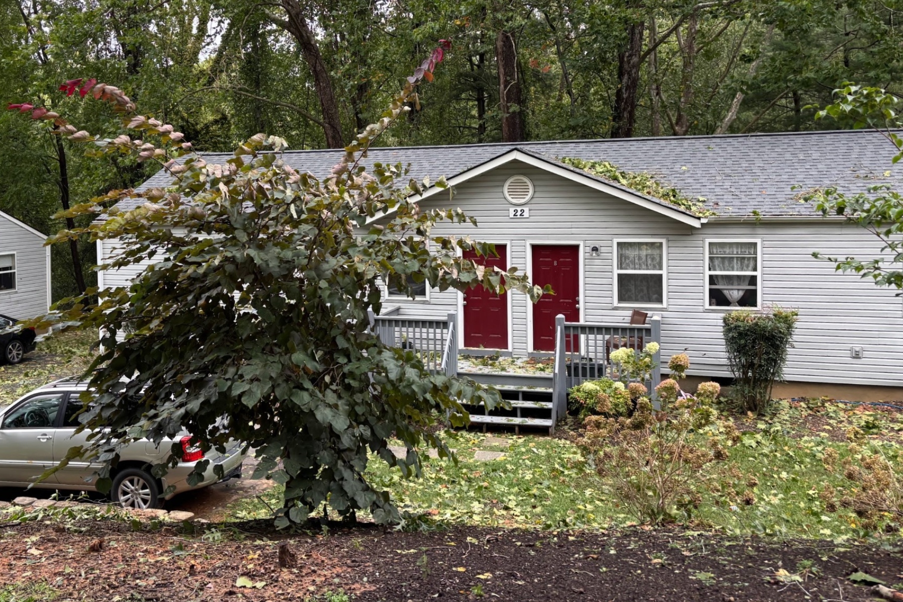 Two red doors on a gray siding duplex with a small front porch, surrounded by trees and parked cars nearby.