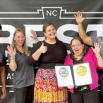 Six smiling coworkers posing with two 2025 winner certificates, raising hands in celebration in front of a dark banner.