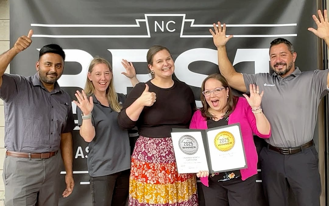 Six smiling coworkers posing with two 2025 winner certificates, raising hands in celebration in front of a dark banner.