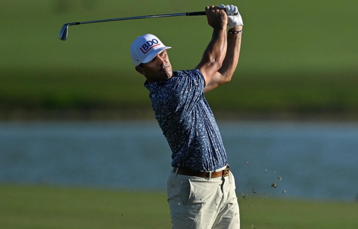 Golfer in a blue patterned shirt mid-swing, on a sunny course with water in the background.