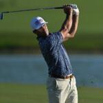 Golfer in a blue patterned shirt mid-swing, on a sunny course with water in the background.