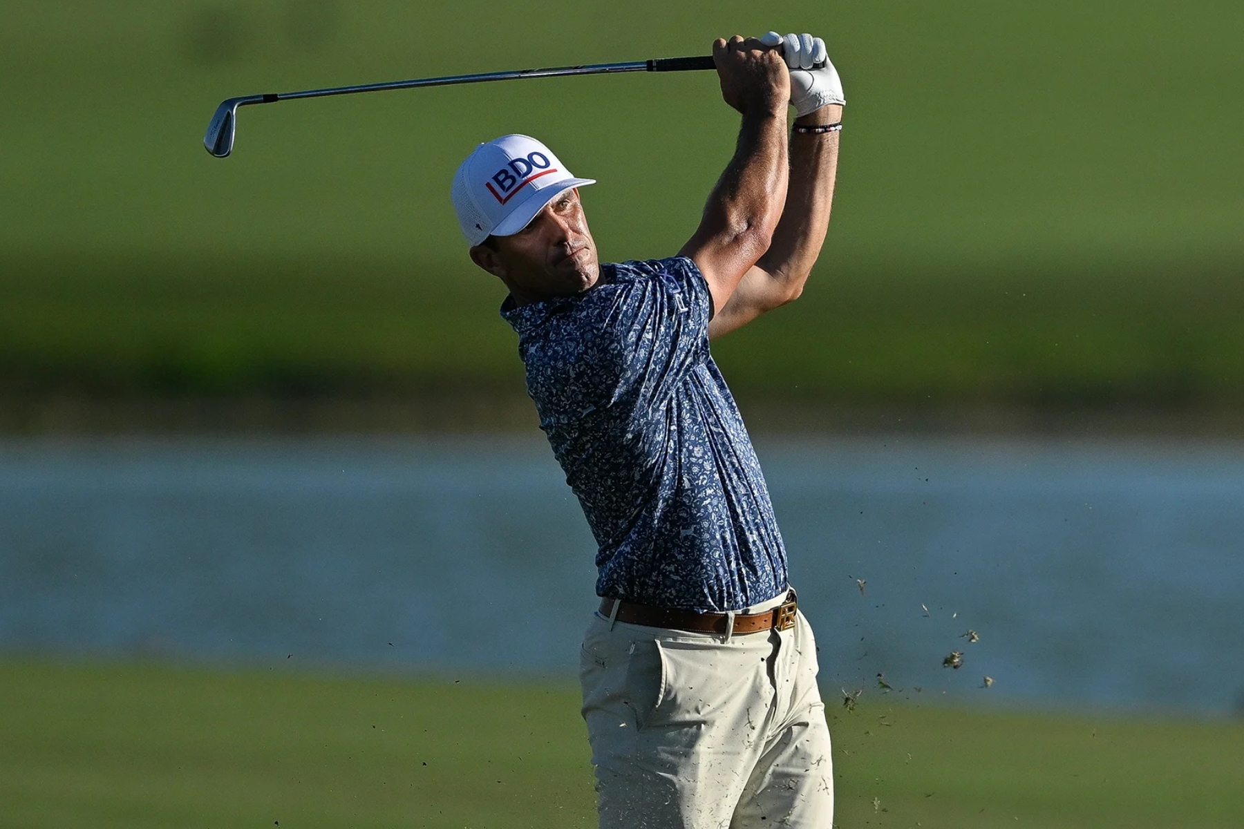 Golfer in a blue patterned shirt mid-swing, on a sunny course with water in the background.