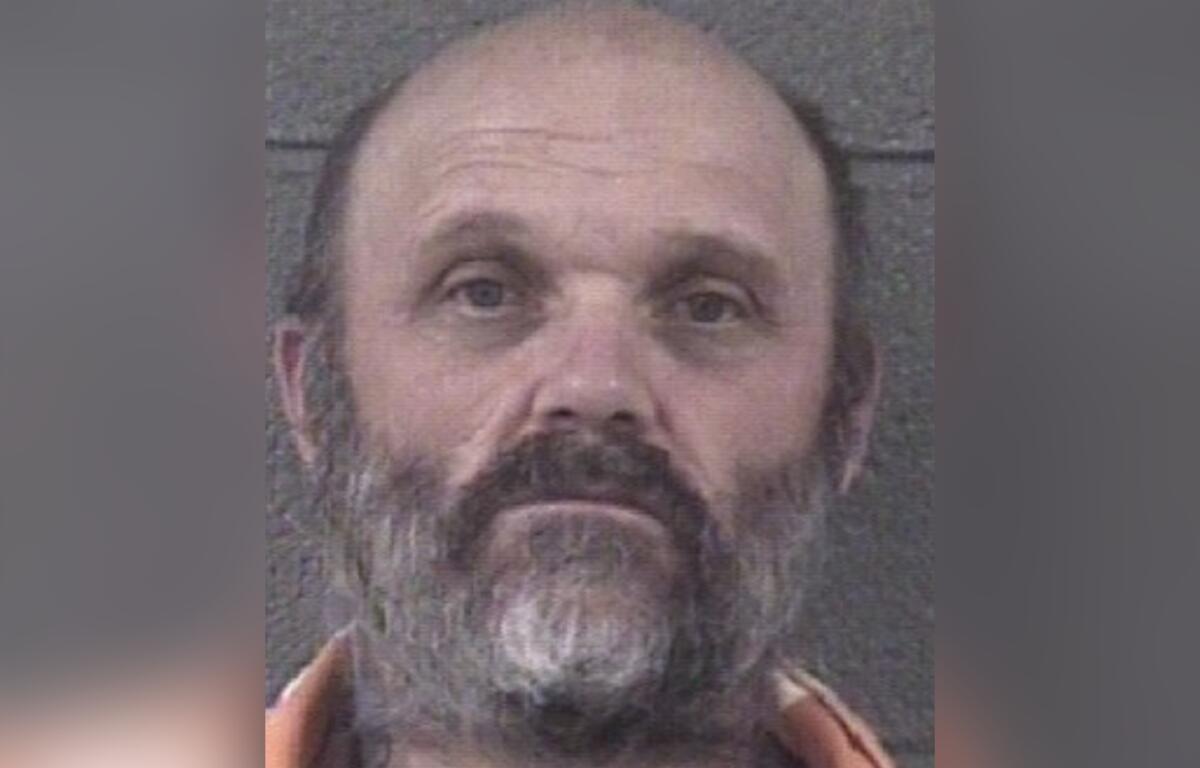 Mugshot of a middle-aged man with a gray beard and mustache, wearing an orange jumpsuit, looking slightly upward.