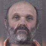 Mugshot of a middle-aged man with a gray beard and mustache, wearing an orange jumpsuit, looking slightly upward.