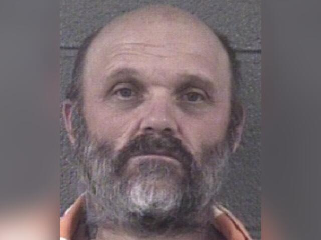 Mugshot of a middle-aged man with a gray beard and mustache, wearing an orange jumpsuit, looking slightly upward.
