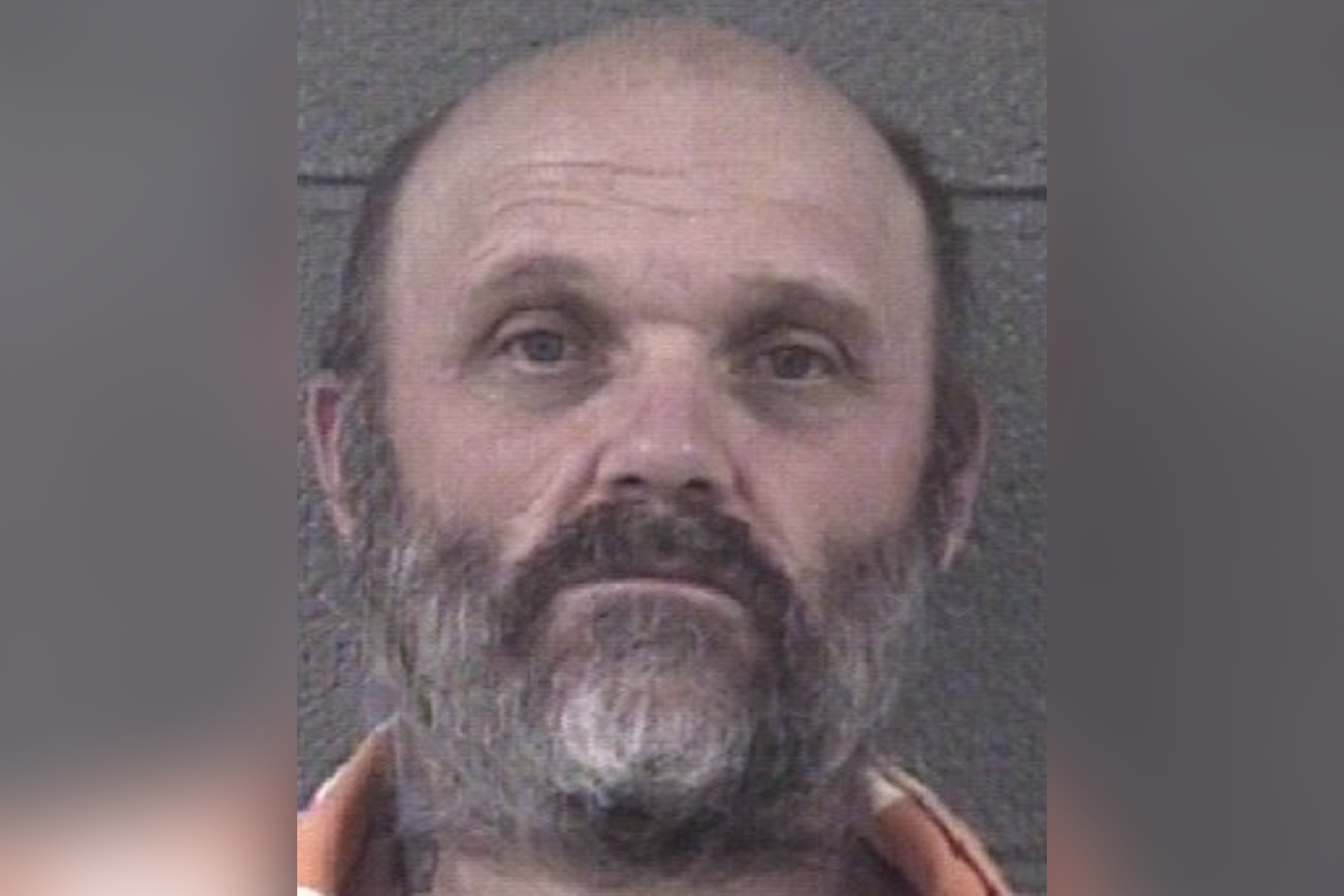 Mugshot of a middle-aged man with a gray beard and mustache, wearing an orange jumpsuit, looking slightly upward.