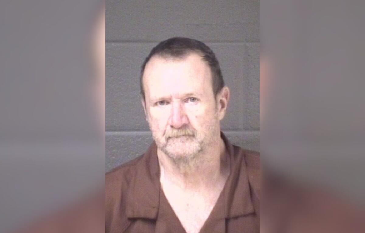 Mugshot of a middle-aged man with short dark hair and a light beard, wearing a brown shirt, against a gray backdrop.