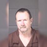 Mugshot of a middle-aged man with short dark hair and a light beard, wearing a brown shirt, against a gray backdrop.
