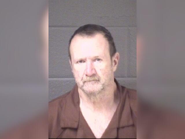 Mugshot of a middle-aged man with short dark hair and a light beard, wearing a brown shirt, against a gray backdrop.