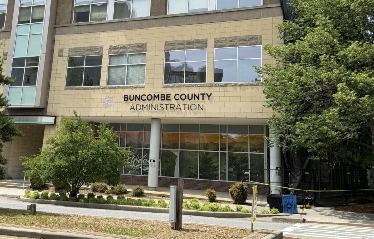 Front of Buncombe County Administration building with beige brick facade and large windows, trees on right, and a pedestrian walkway in front.