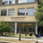 Front of Buncombe County Administration building with beige brick facade and large windows, trees on right, and a pedestrian walkway in front.
