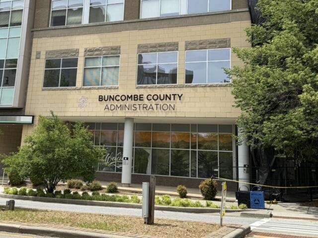 Front of Buncombe County Administration building with beige brick facade and large windows, trees on right, and a pedestrian walkway in front.