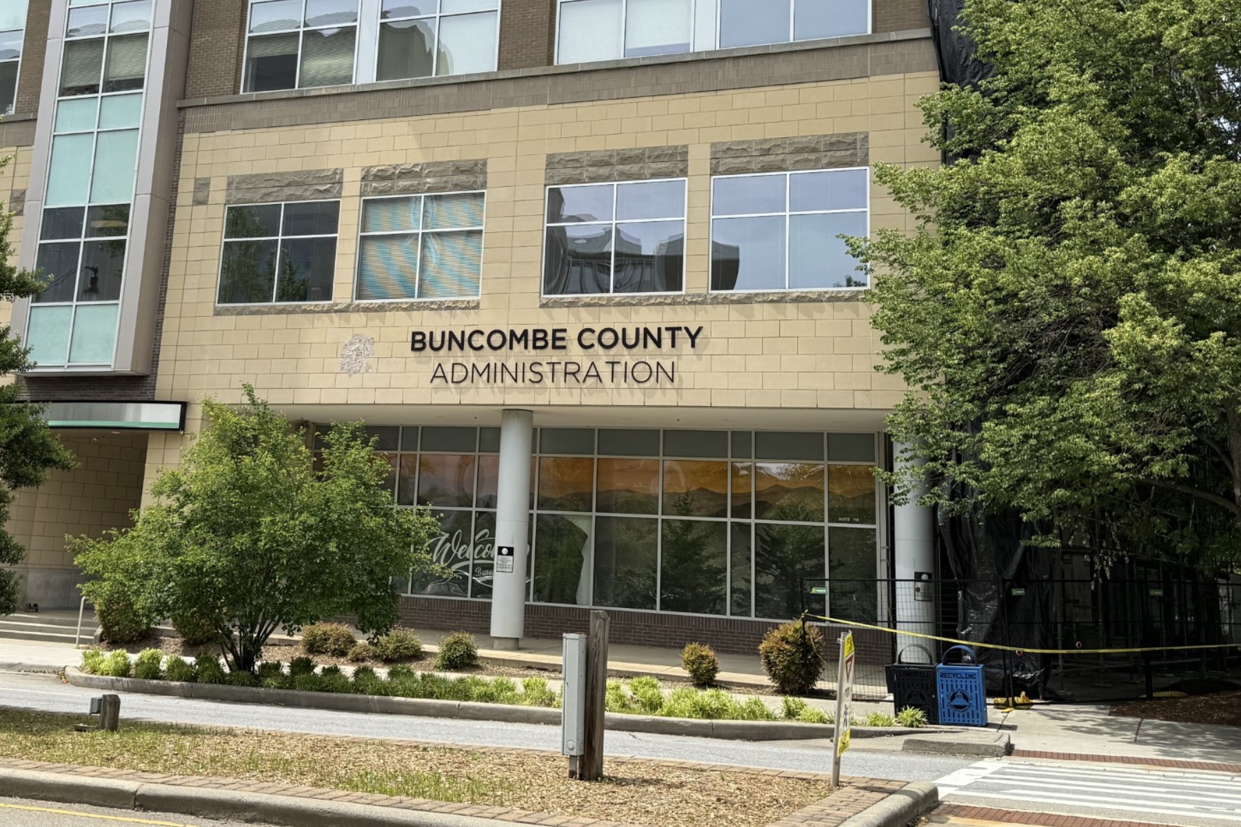 Front of Buncombe County Administration building with beige brick facade and large windows, trees on right, and a pedestrian walkway in front.