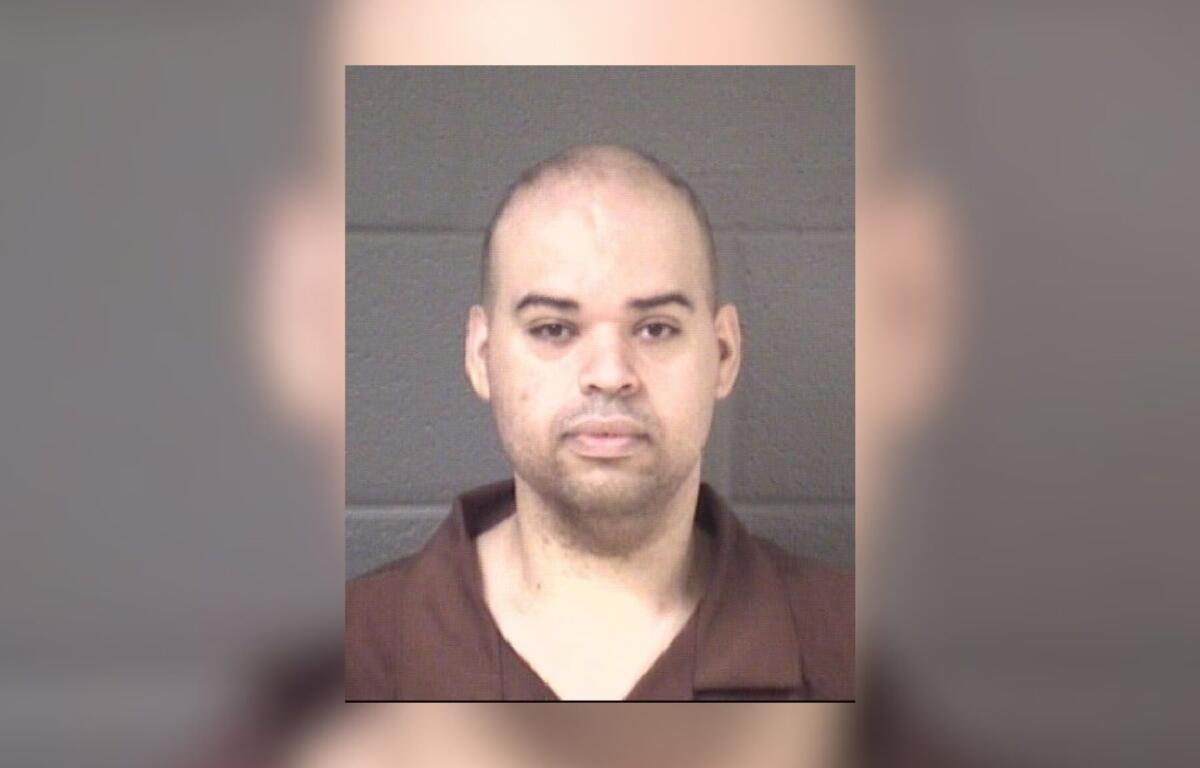 Mugshot of a man with short hair in a brown shirt against a gray wall.