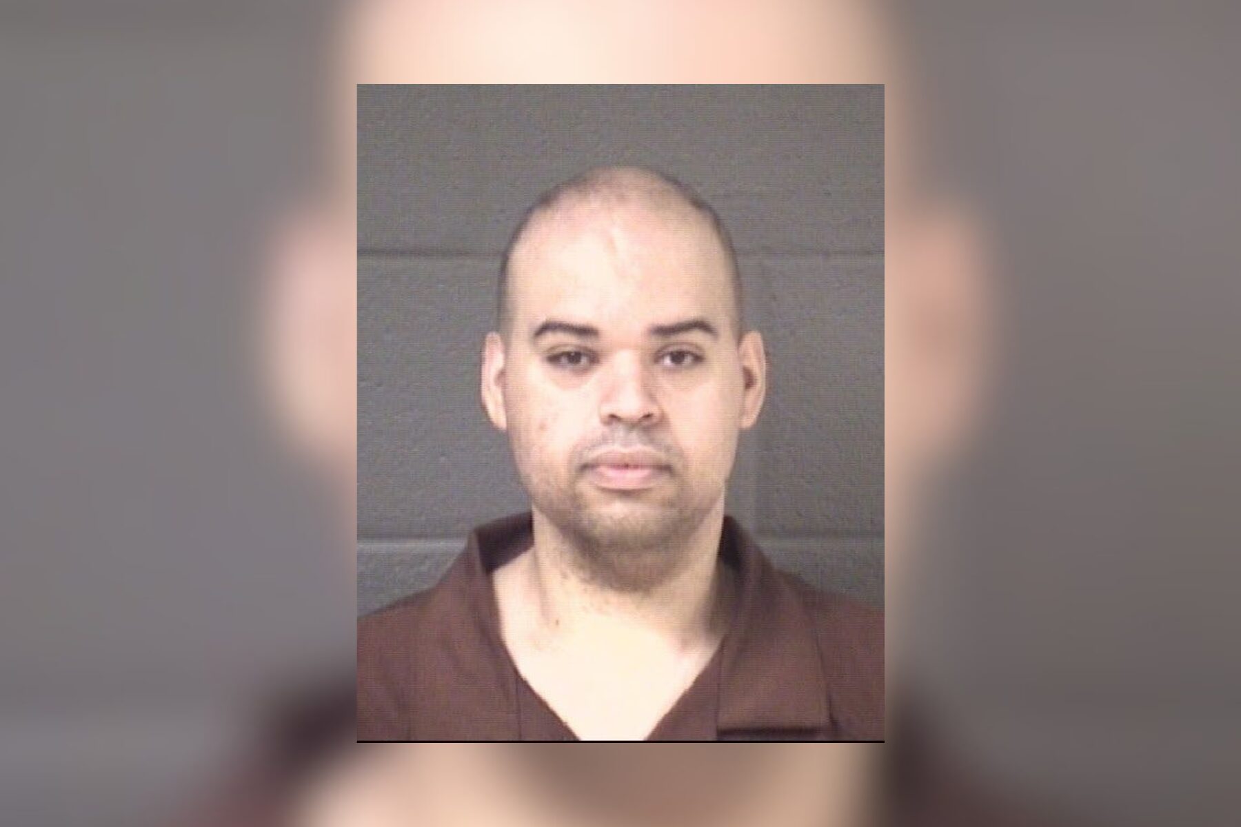 Mugshot of a man with short hair in a brown shirt against a gray wall.