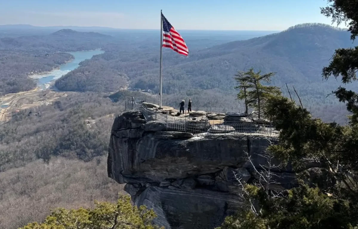 American flag on a tall pole at a cliff viewpoint, two people standing near a railing with a panoramic valley below.