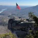 American flag on a tall pole at a cliff viewpoint, two people standing near a railing with a panoramic valley below.