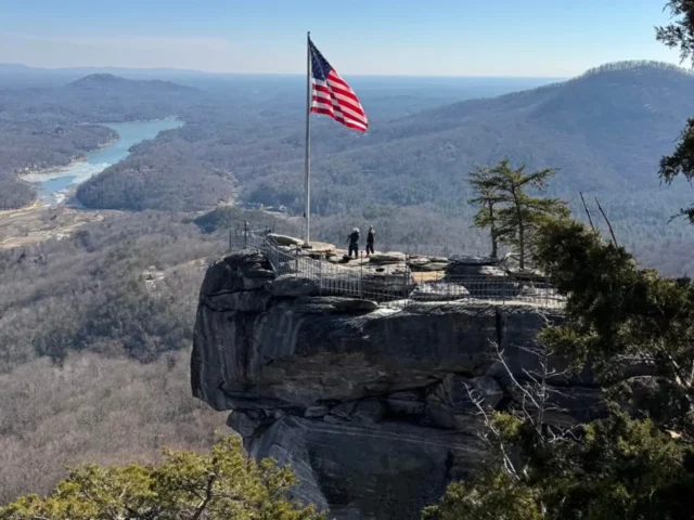 American flag on a tall pole at a cliff viewpoint, two people standing near a railing with a panoramic valley below.