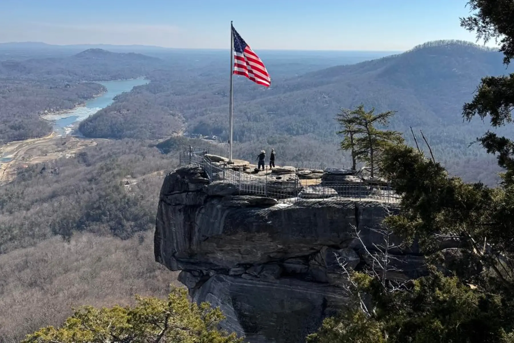 American flag on a tall pole at a cliff viewpoint, two people standing near a railing with a panoramic valley below.