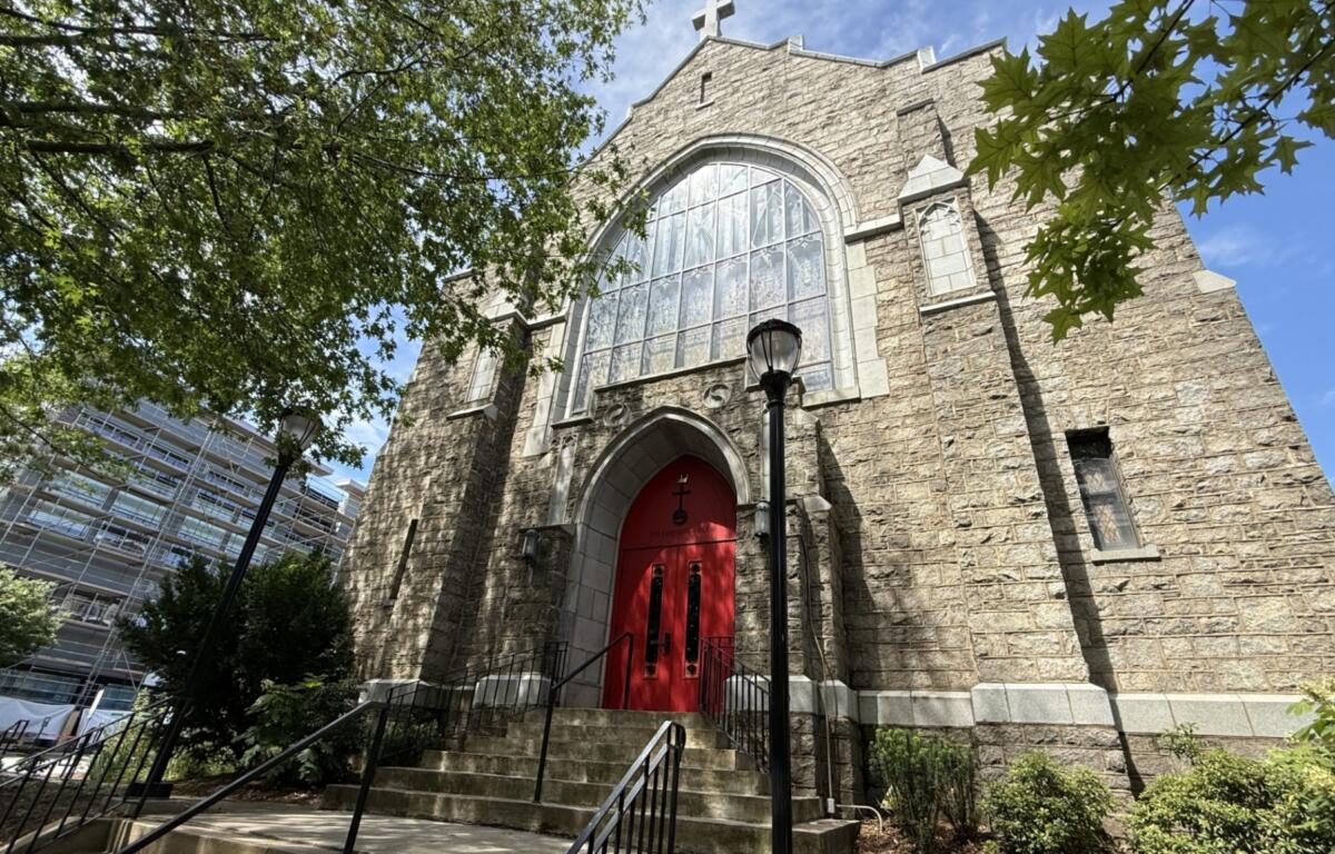 Stone church with a large arched window and red doors, framed by green trees against a blue sky, on a sunny day.