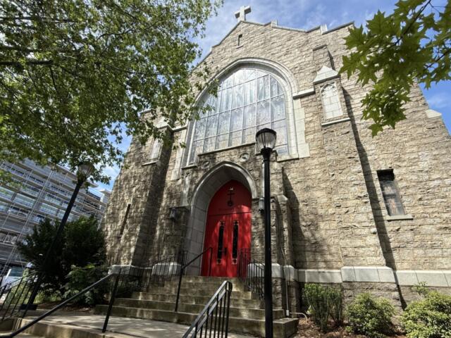 Stone church with a large arched window and red doors, framed by green trees against a blue sky, on a sunny day.