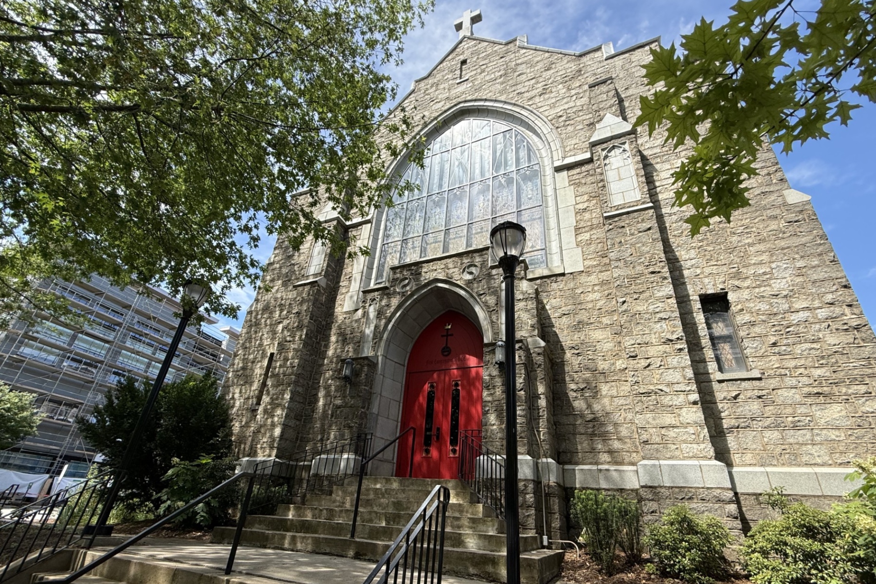 Stone church with a large arched window and red doors, framed by green trees against a blue sky, on a sunny day.