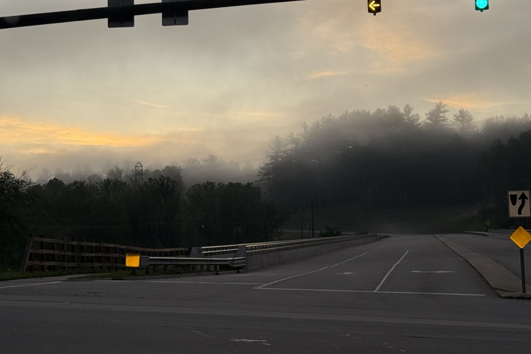 Morning fog over a road with guardrails, trees in the distance, and a green traffic light overhead on the right.