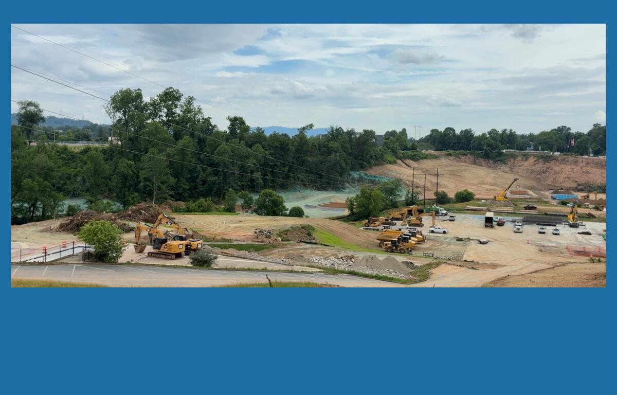 Construction site with excavators, trucks, and a dirt hill beside a forest under a cloudy sky on a sunny day, with power lines above.