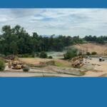 Construction site with excavators, trucks, and a dirt hill beside a forest under a cloudy sky on a sunny day, with power lines above.
