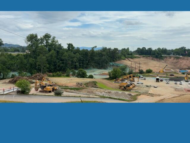 Construction site with excavators, trucks, and a dirt hill beside a forest under a cloudy sky on a sunny day, with power lines above.