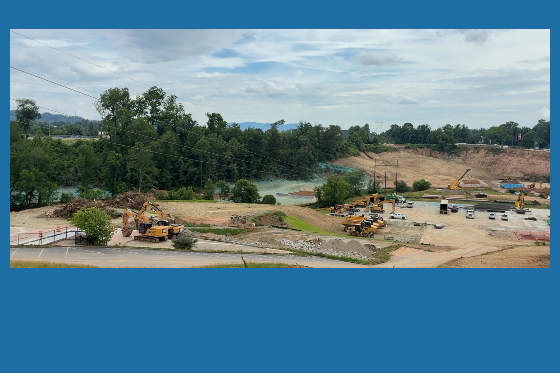 Construction site with excavators, trucks, and a dirt hill beside a forest under a cloudy sky on a sunny day, with power lines above.