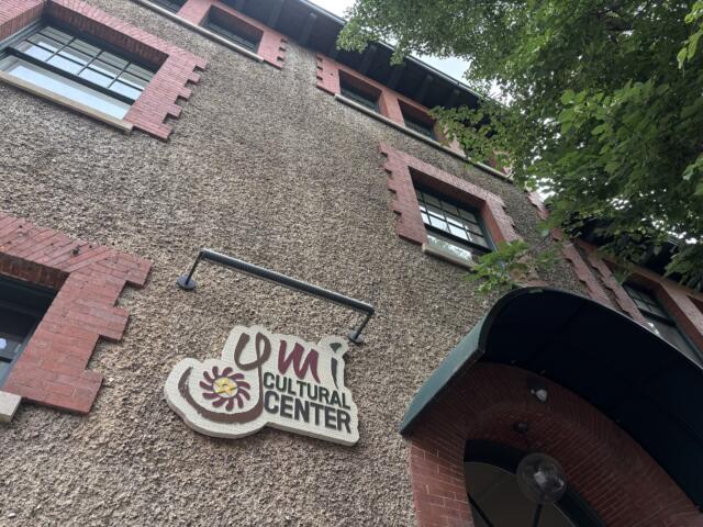 Exterior of a textured beige pebble-dash building with red brick window trim and a Cultural Center sign, with leafy branches on the right.