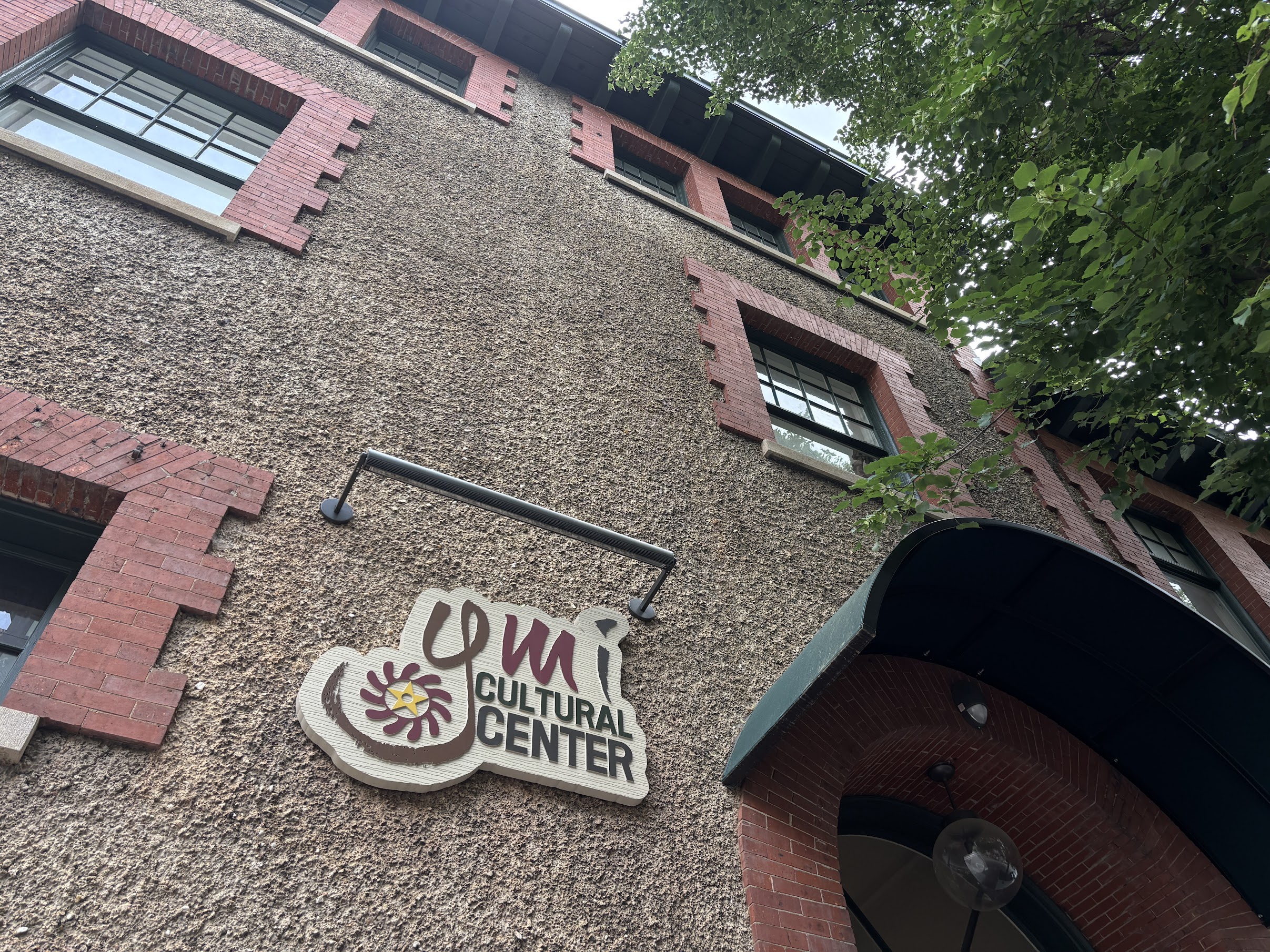 Exterior of a textured beige pebble-dash building with red brick window trim and a Cultural Center sign, with leafy branches on the right.
