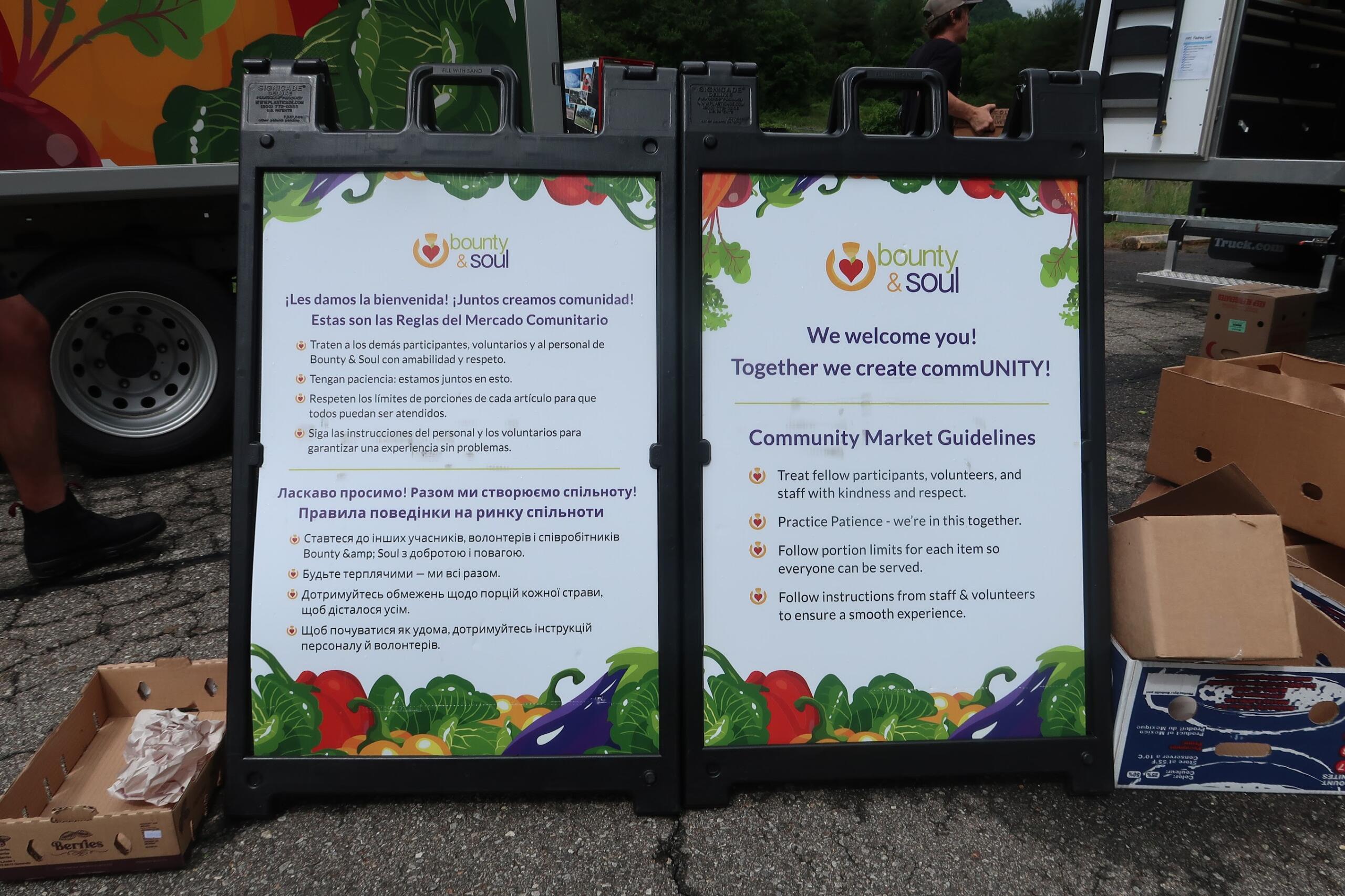 Informational sign panels for a community market (Bounty & Soul) with colorful borders and multilingual guidelines, outdoors by a truck.