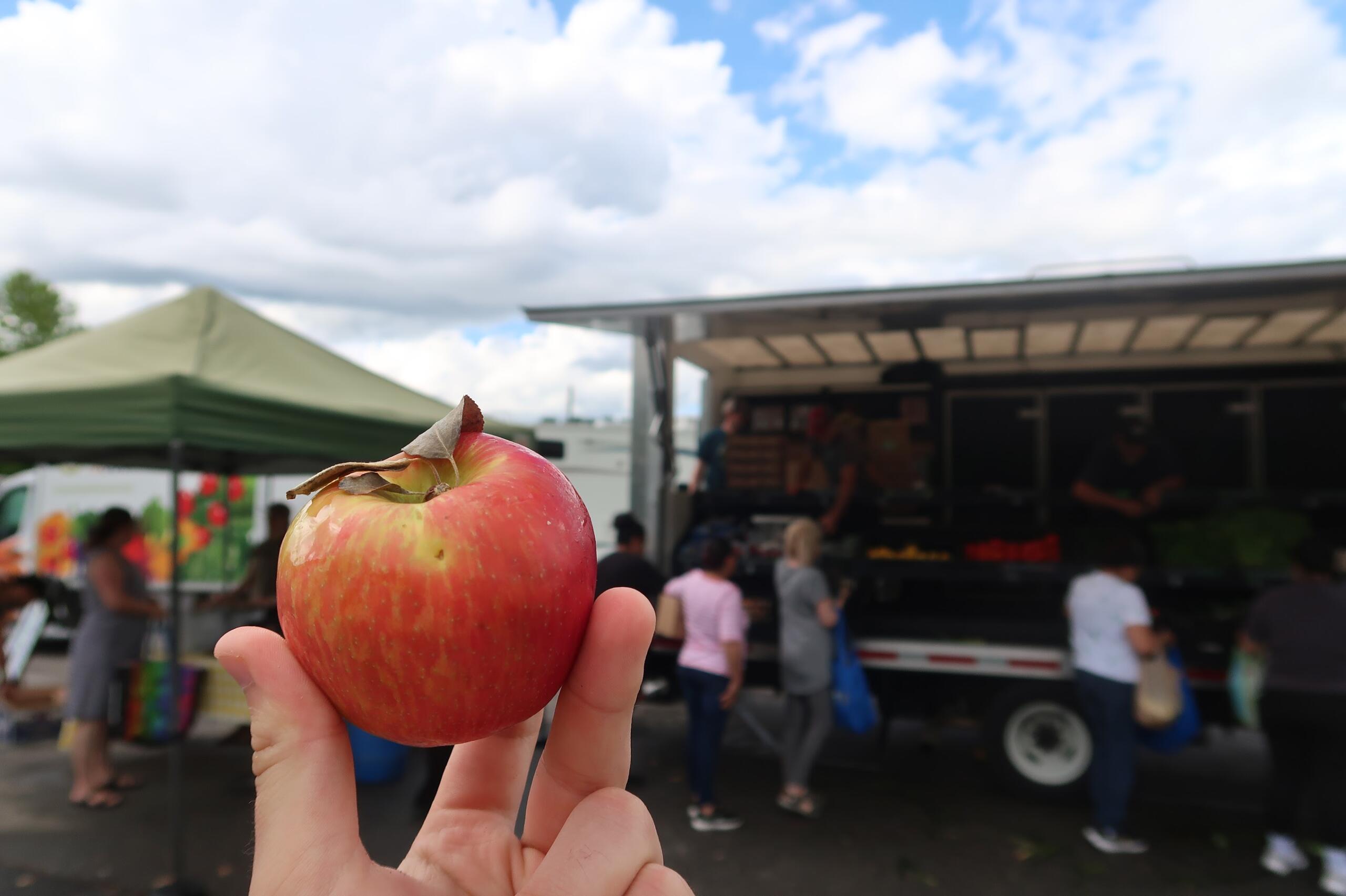 Close-up of a red apple held between fingers at a busy outdoor market.