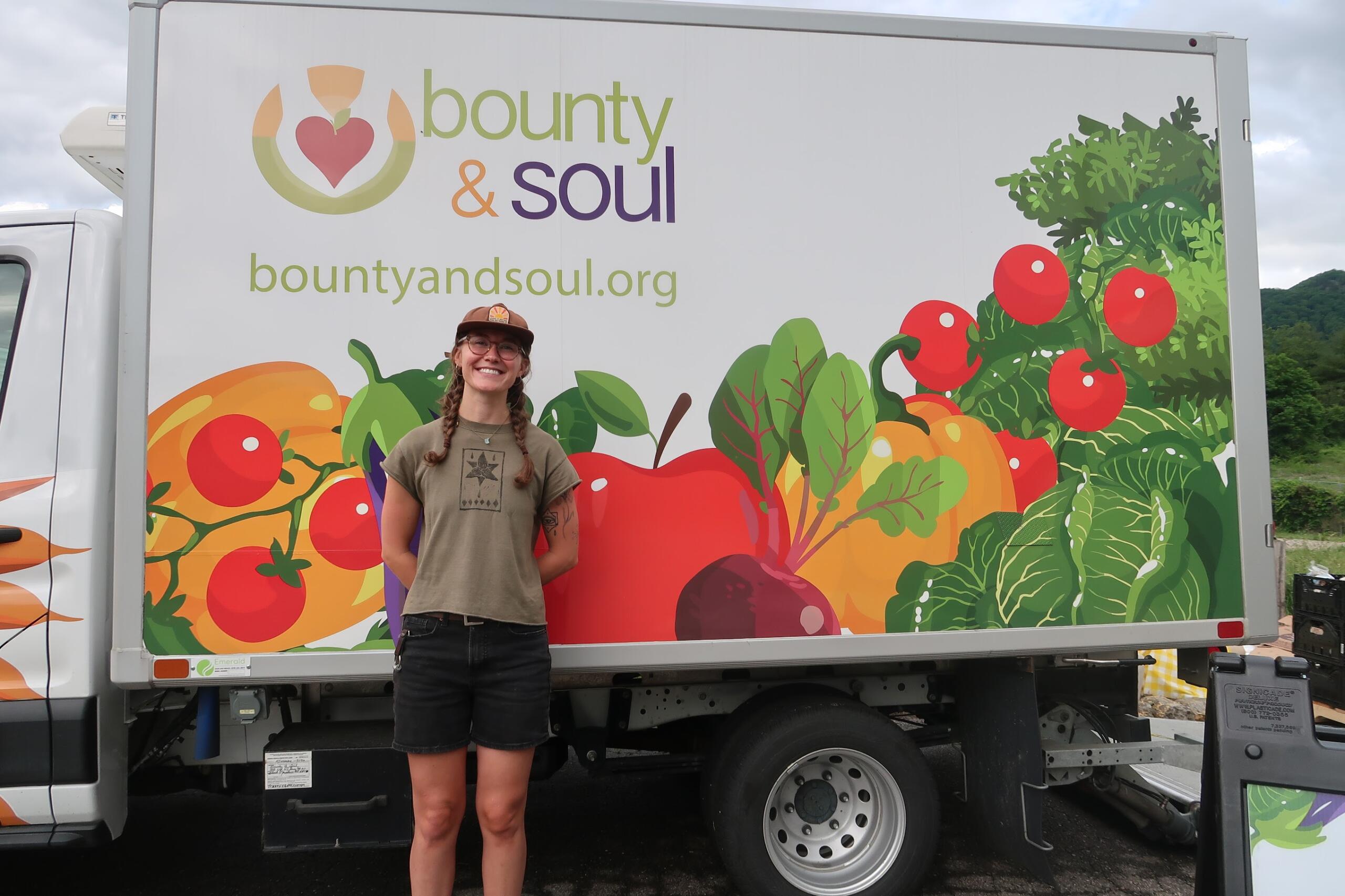Person with braids and a brown cap standing in front of a colorful 'bounty & soul' truck mural featuring vegetables and fruit.