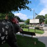 Life-size cow statue painted with white letters in a park, with the Museum of the Cherokee People sign visible across the street.